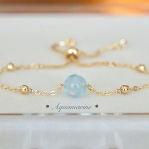 Puede incluir: Delicada pulsera dorada con una piedra central de aguamarina azul claro. La pulsera tiene un diseño de cadena fina con pequeñas cuentas doradas. La palabra "Aquamarine" está impresa debajo.