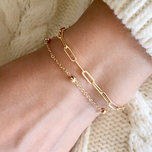 May include: Two gold bracelets on a wrist. One bracelet is a delicate chain with small round beads. The other bracelet is a thicker chain with larger oval links.