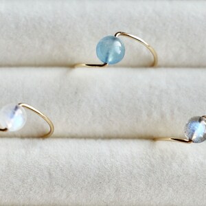 May include: Three gold rings with different colored gemstones. The rings are made of thin gold wire and have a simple design. The gemstones are a light blue aquamarine, a white moonstone, and a grey labradorite.