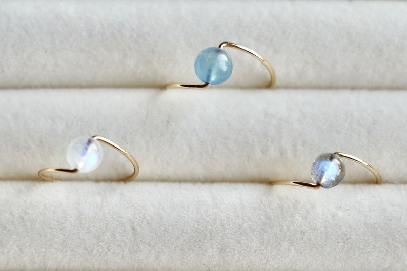 May include: Three gold rings with different colored gemstones. The rings are made of thin gold wire and have a simple design. The gemstones are a light blue aquamarine, a white moonstone, and a grey labradorite.
