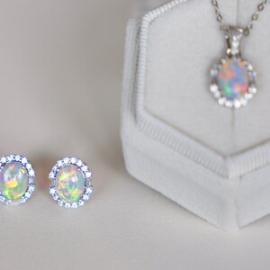 May include: A set of opal jewelry featuring a pendant necklace and a pair of earrings. The necklace and earrings have a silver setting with a halo of small clear stones surrounding the opal.
