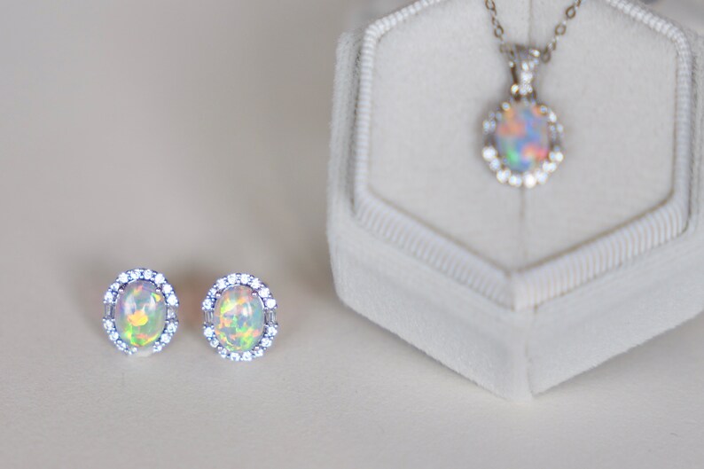 May include: A set of opal jewelry featuring a pendant necklace and a pair of earrings. The necklace and earrings have a silver setting with a halo of small clear stones surrounding the opal.