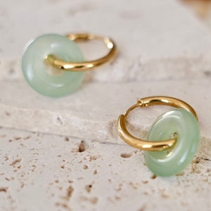 May include: A pair of gold hoop earrings with a light green jade stone accent. The jade stone is shaped like a donut and is attached to the hoop with a gold ring.