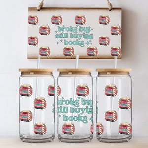 Può includere: Tre bicchieri di vetro trasparente con coperchi in bambù e cannucce, ognuno decorato con una grafica di pila di libri. Un cartello di legno sopra ripete la grafica e il testo "broke but still buying books."