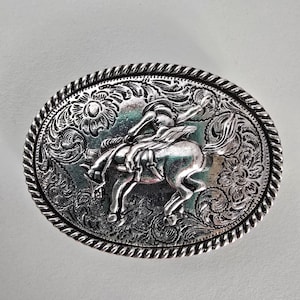 May include: Oval silver-coloured belt buckle depicting a cowboy riding a bucking bronco. The buckle has an intricate floral and sunburst design around the edges and the cowboy and horse. The border has a rope-like texture.