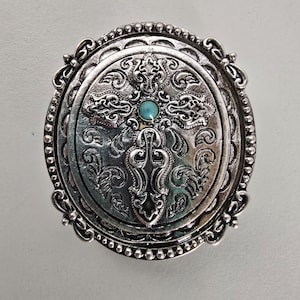 May include: Silver-toned, round belt buckle with an ornate cross design. The center features a small, turquoise-colored stone. The buckle has a detailed, embossed pattern and a beaded border, showcasing a vintage or Western aesthetic.