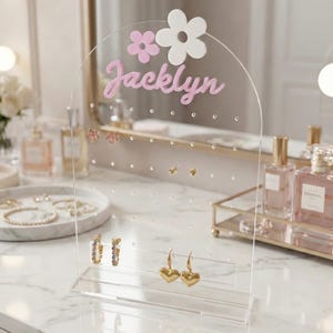 Op de afbeelding: Transparante acryl oorbellenhouder met de naam "Jacklyn" in roze letters, versierd met roze en witte bloemen. Het heeft meerdere gaten voor oorbellen en een basis voor het ophangen van oorbellen. Gouden hartvormige oorbellen worden getoond.