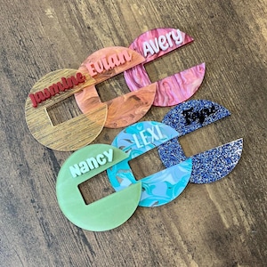 Peut inclure: Plusieurs étiquettes nominatives personnalisées en acrylique, aux couleurs et motifs variés. Chaque étiquette présente un nom différent dans une couleur contrastante, dont "Jasmine", "Eutan", "Avery", "Lexi", "Nancy", et "Taya". Les étiquettes sont circulaires avec une découpe rectangulaire.