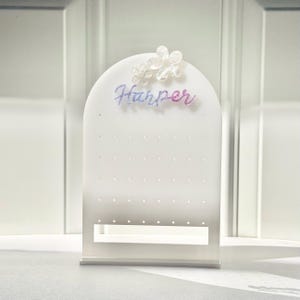 May include: A white arched acrylic earring display stand featuring a grid of small holes for jewelry organization. The top is decorated with two translucent flower shapes and the name Harper in a purple and blue gradient script font.