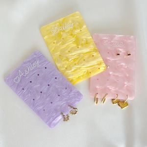 May include: Three rectangular acrylic earring display cards in lavender, yellow, and pink with a marbled finish. Each card features a custom name engraved at the top: Ashley, Bailey, and Kaia. The cards have multiple holes for jewelry.