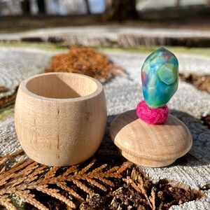 Puede incluir: Una pequeña caja de madera con tapa. La tapa está coronada con un círculo de fieltro rosa y una piedra iridiscente azul y verde. La caja está sobre una superficie de madera.