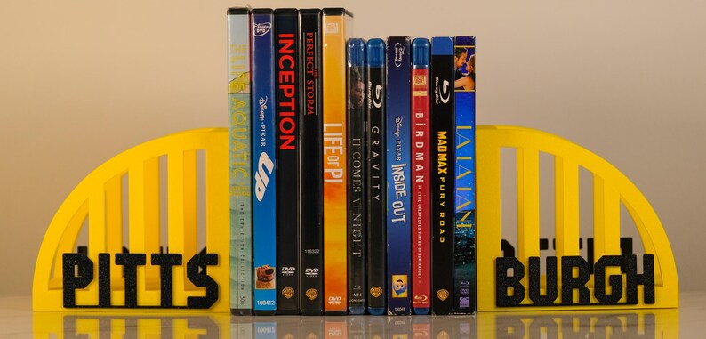 Puede incluir: Una colecci&oacute;n de DVDs y Blu-ray se mantiene entre sujetalibros amarillos con la palabra "PITTSBURGH" en letras negras. Los t&iacute;tulos de las pel&iacute;culas incluyen "Up", "Inception" y "Mad Max: Fury Road".