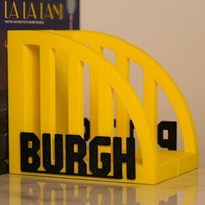 Puede incluir: Un sujetalibros amarillo brillante con la palabra "BURGH" en letras negras. El soporte tiene un dise&ntilde;o similar a un puente con soportes arqueados. Dise&ntilde;ado para guardar libros y revistas.