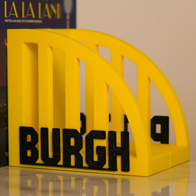 Puede incluir: Un sujetalibros amarillo brillante con la palabra "BURGH" en letras negras. El soporte tiene un dise&ntilde;o similar a un puente con soportes arqueados. Dise&ntilde;ado para guardar libros y revistas.