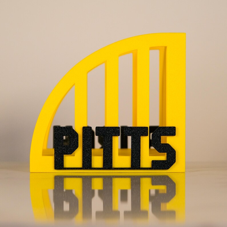 Puede incluir: Un organizador de escritorio decorativo amarillo y negro con la palabra "PITTS" en letras negras texturizadas. El organizador tiene un dise&ntilde;o curvo y arqueado con barras verticales. Las letras se reflejan en la superficie inferior.