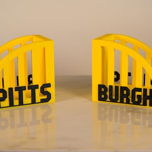 Puede incluir: Dos sujetalibros amarillos en forma de arco con letras negras que deletrean "PITTS" y "BURGH". Los sujetalibros tienen un dise&ntilde;o ranurado y se muestran sobre una superficie reflectante. El tema general es la decoraci&oacute;n del hogar.