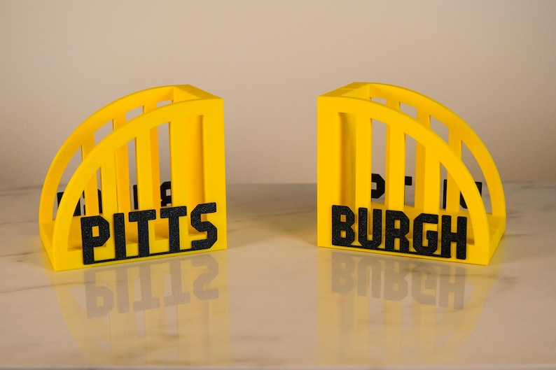 Puede incluir: Dos sujetalibros amarillos en forma de arco con letras negras que deletrean "PITTS" y "BURGH". Los sujetalibros tienen un dise&ntilde;o ranurado y se muestran sobre una superficie reflectante. El tema general es la decoraci&oacute;n del hogar.
