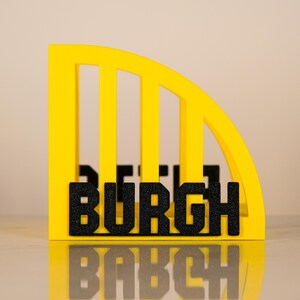 Puede incluir: Un letrero decorativo amarillo y negro con la palabra "BURGH" en letras negras y brillantes. El letrero tiene un dise&ntilde;o arqueado con barras verticales, y la palabra "BURGH" se muestra de forma destacada.