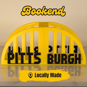 Puede incluir: Un sujetalibros amarillo y negro con forma de puente, con la palabra "Bookend" en escritura amarilla. Presenta la palabra "PITTSBURGH" en letras negras. Tambi&eacute;n est&aacute; presente el texto "Locally Made".