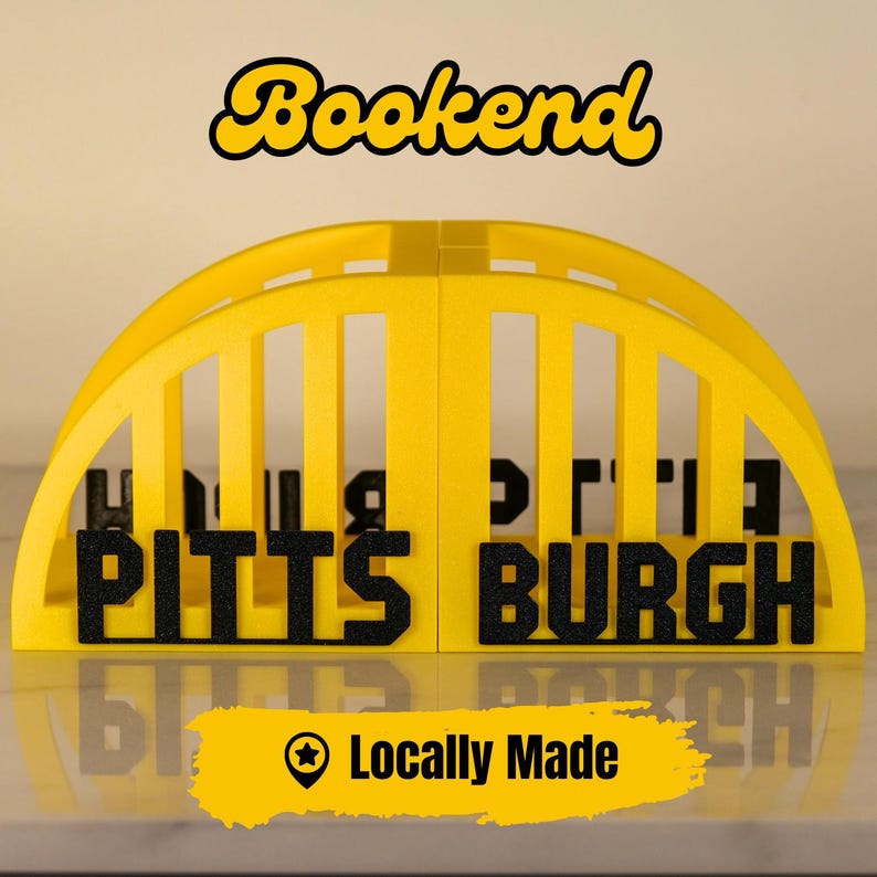Puede incluir: Un sujetalibros amarillo y negro con forma de puente, con la palabra "Bookend" en escritura amarilla. Presenta la palabra "PITTSBURGH" en letras negras. Tambi&eacute;n est&aacute; presente el texto "Locally Made".
