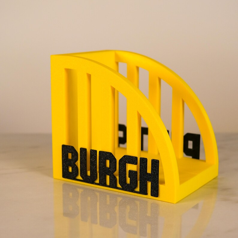 Puede incluir: Un revistero amarillo brillante con una parte superior curvada y divisores verticales. La palabra "BURGH" se muestra en letras negras texturizadas en la parte delantera. Dise&ntilde;ado para guardar revistas o archivos.