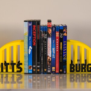 Puede incluir: Un juego de sujetalibros amarillos con la palabra "Pittsburgh" en negro. Soportan una colecci&oacute;n de DVD y Blu-ray, incluyendo "Up", "Inception" y "La La Land".