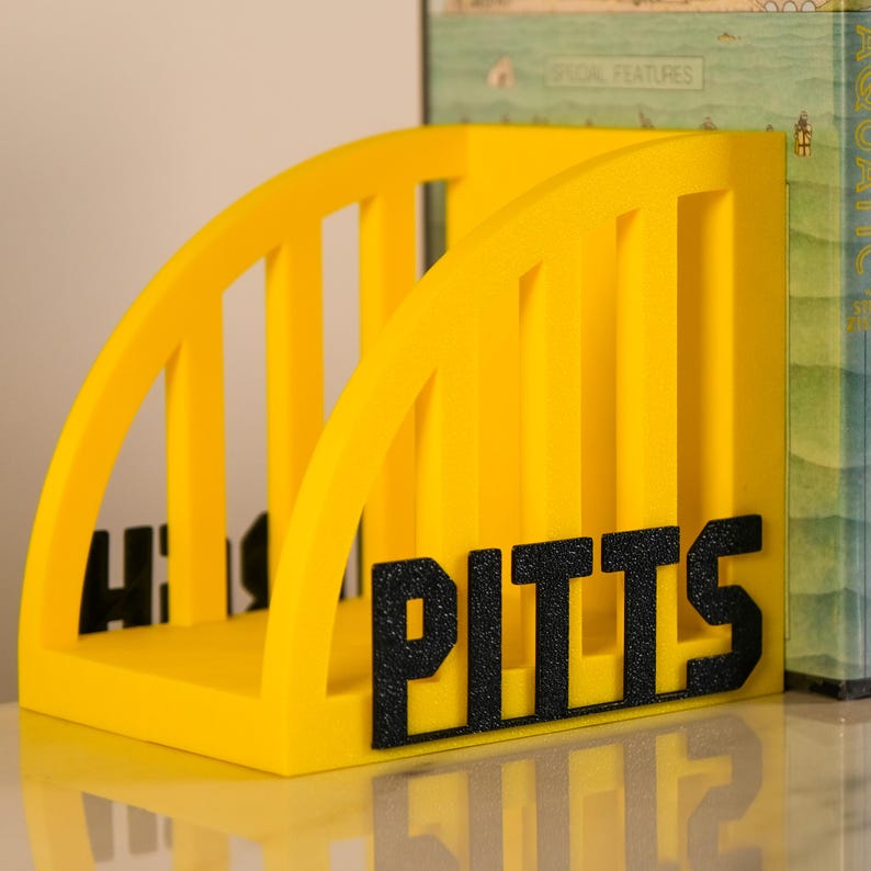 Puede incluir: Un sujetalibros amarillo brillante con la palabra "PITTS" en letras negras en negrita. El sujetalibros tiene un dise&ntilde;o arqueado con soportes verticales. Las letras est&aacute;n en relieve, creando una apariencia texturizada. Dise&ntilde;ado para sujetar libros.