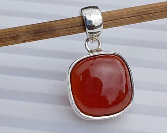 May include: A square, reddish-orange pendant with a polished surface, set in a silver-colored metal frame. The pendant hangs from a silver bail. The background is a light-colored surface.