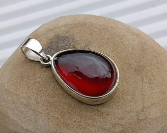 May include: A teardrop-shaped pendant with a deep red gemstone set in a silver bezel. The gemstone has a gradient effect, transitioning from dark to light red. The pendant has a silver loop for a chain.