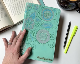 May include: A teal journal with embossed floral designs in gray and blue. The journal has the name "Marilyn Hess" printed in gold. A black pen and a yellow highlighter are next to the journal. A cup of coffee and an open book are also in the image.