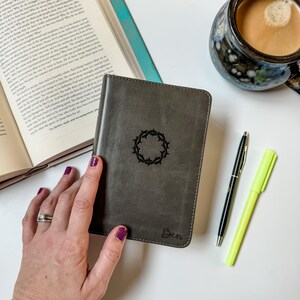 May include: Gray leather journal with a black crown of thorns design and the name "Ben" embossed on the cover. A black pen and a yellow highlighter are next to the journal. A cup of coffee and an open book are in the background.