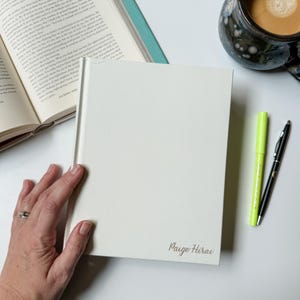 May include: A white hardcover journal with the name "Paige Hirai" written in gold lettering on the front cover. The journal is lying on a white surface with a green highlighter and a black pen next to it.