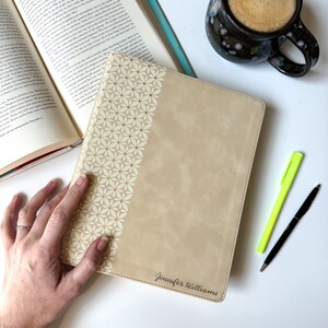 May include: Tan Bible cover with a floral design on the left side and the name "Jennifer Williams" at the bottom. A cup of coffee, two pens, and an open book are also visible.
