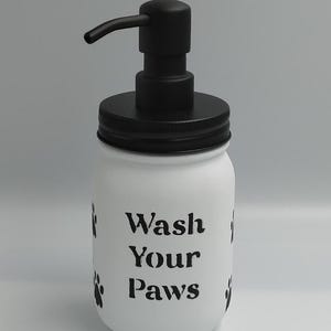 Puede incluir: Dispensador de jabón de tarro Mason blanco con bomba y tapa negras. El tarro está decorado con huellas de patas negras y las palabras "Wash Your Paws". Un artículo decorativo y funcional para el baño o la cocina.