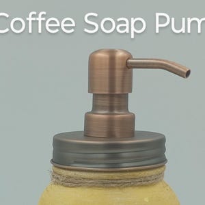 May include: A close-up of a coffee soap pump. The pump is made of copper-colored metal and is attached to a jar with a yellow base. The words "Coffee Soap Pump" are written in white letters at the top.