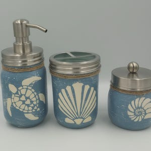 Puede incluir: Un juego de tres accesorios de baño con temática costera. El dispensador de jabón, el portacepillos de dientes y el tarro de bastoncillos de algodón son todos de vidrio con un fondo azul y diseños marinos blancos. El dispensador de jabón tiene una bomba plateada y el portacepillos de dientes tiene una tapa plateada.
