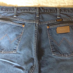 Puede incluir: Primer plano de la parte trasera de unos vaqueros azules. Los vaqueros tienen dos bolsillos traseros con una costura en forma de 'W' en hilo naranja. Una etiqueta de cuero marr&oacute;n con el nombre de la marca es visible en el bolsillo derecho. Remaches de cobre.