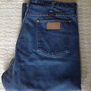 Puede incluir: Vaqueros de mezclilla azul oscuro con un dise&ntilde;o cl&aacute;sico de cinco bolsillos. Los vaqueros tienen un parche de cuero marr&oacute;n con el logotipo "Wrangler" en el bolsillo trasero. Los vaqueros est&aacute;n doblados.