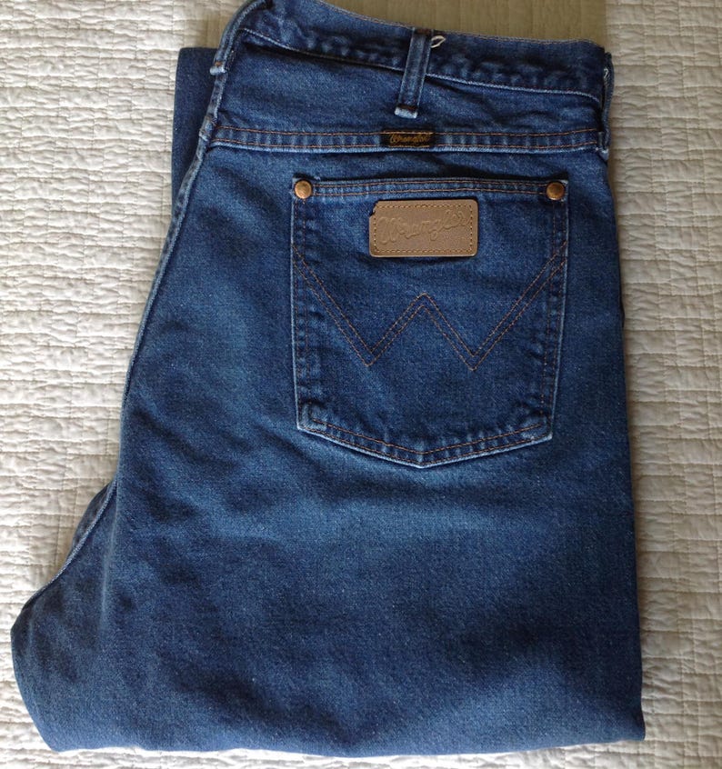 Puede incluir: Vaqueros de mezclilla azul oscuro con un dise&ntilde;o cl&aacute;sico de cinco bolsillos. Los vaqueros tienen un parche de cuero marr&oacute;n con el logotipo "Wrangler" en el bolsillo trasero. Los vaqueros est&aacute;n doblados.