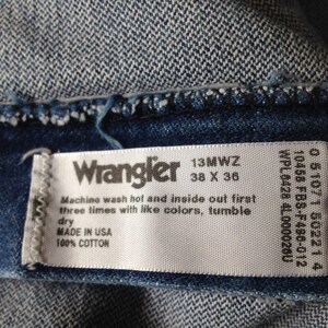 Puede incluir: Primer plano de una prenda vaquera con una etiqueta blanca. La etiqueta dice "Wrangler 13MWZ 38 x 36" e incluye instrucciones de lavado. Fabricado en EE. UU. con 100% algod&oacute;n. El vaquero es de color azul medio.