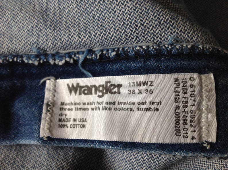 Puede incluir: Primer plano de una prenda vaquera con una etiqueta blanca. La etiqueta dice "Wrangler 13MWZ 38 x 36" e incluye instrucciones de lavado. Fabricado en EE. UU. con 100% algod&oacute;n. El vaquero es de color azul medio.