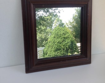 May include: A square, dark brown framed mirror. The mirror reflects a green tree and other foliage. The frame has a detailed, ridged design. The mirror is set on a white surface.