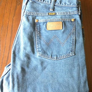 Puede incluir: Vaqueros de mezclilla azul claro con un dise&ntilde;o cl&aacute;sico de cinco bolsillos. Los vaqueros presentan un parche de cuero con el logotipo "Wrangler" en el bolsillo trasero y una peque&ntilde;a etiqueta met&aacute;lica encima del bolsillo trasero. Los vaqueros est&aacute;n doblados cuidadosamente.