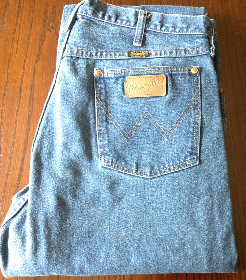 Puede incluir: Vaqueros de mezclilla azul claro con un dise&ntilde;o cl&aacute;sico de cinco bolsillos. Los vaqueros presentan un parche de cuero con el logotipo "Wrangler" en el bolsillo trasero y una peque&ntilde;a etiqueta met&aacute;lica encima del bolsillo trasero. Los vaqueros est&aacute;n doblados cuidadosamente.