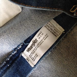 Puede incluir: Primer plano del interior de unos vaqueros Wrangler. Una etiqueta blanca muestra la marca, la talla 38 x 36 pulgadas e instrucciones de lavado. Los vaqueros son 100% algod&oacute;n y est&aacute;n fabricados en EE. UU.