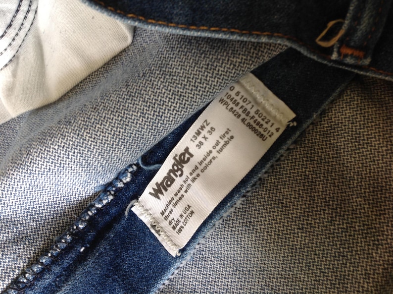 Puede incluir: Primer plano del interior de unos vaqueros Wrangler. Una etiqueta blanca muestra la marca, la talla 38 x 36 pulgadas e instrucciones de lavado. Los vaqueros son 100% algod&oacute;n y est&aacute;n fabricados en EE. UU.
