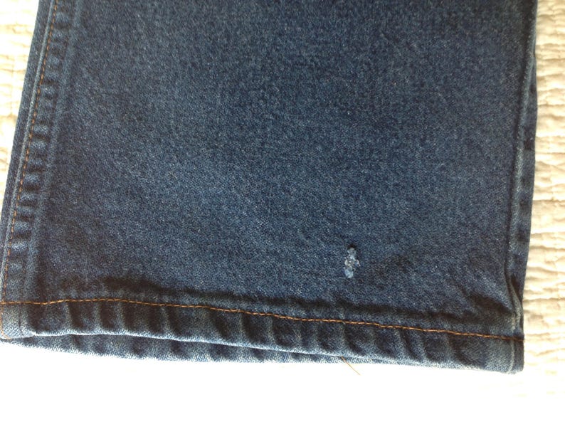 Puede incluir: Primer plano de una secci&oacute;n de vaqueros azul oscuro. La tela tiene un aspecto texturizado con una peque&ntilde;a mancha de color claro. El dobladillo es visible, con detalles de costura marr&oacute;n.