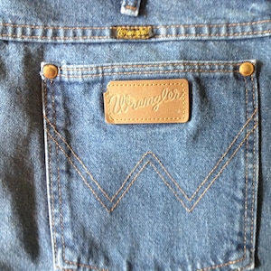Puede incluir: Primer plano de unos vaqueros azules con un bolsillo trasero con el logotipo de Wrangler. El bolsillo tiene un dise&ntilde;o de 'W' cosido y un parche de cuero marr&oacute;n. Los vaqueros tienen remaches y costuras de color cobre.