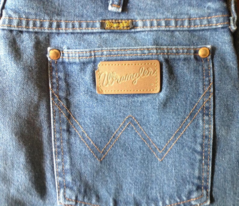 Puede incluir: Primer plano de unos vaqueros azules con un bolsillo trasero con el logotipo de Wrangler. El bolsillo tiene un dise&ntilde;o de 'W' cosido y un parche de cuero marr&oacute;n. Los vaqueros tienen remaches y costuras de color cobre.
