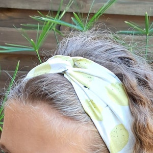 Puede incluir: Una diadema blanca y amarilla con un diseño de limones. La diadema está anudada en la parte superior y se lleva en la cabeza. El fondo incluye una valla de madera y plantas verdes.